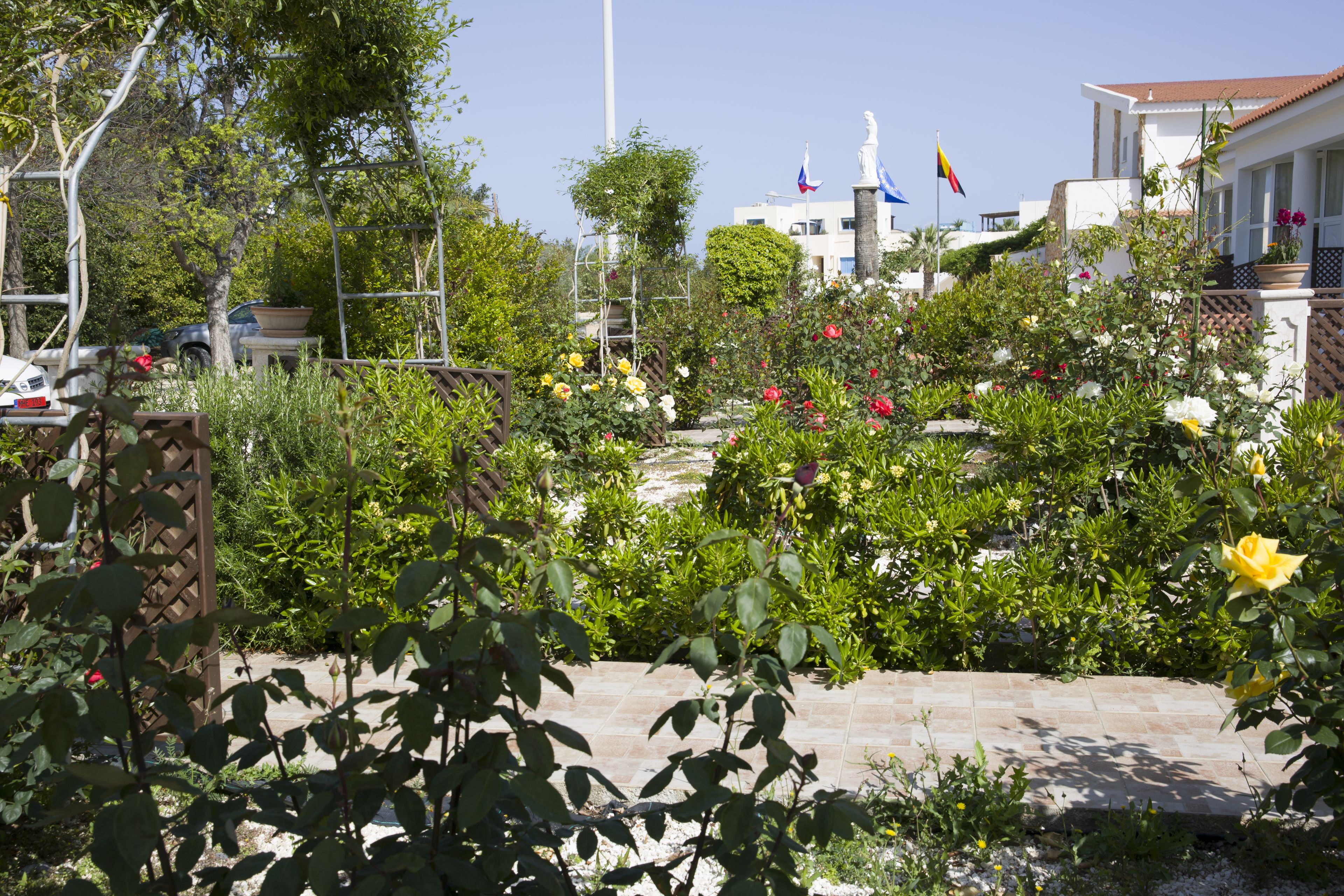 A garden with various plants and flowers, a statue, and flags on a building in the background.