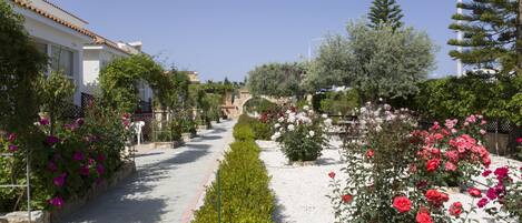 A pathway lined with neatly trimmed hedges and flowering plants, leading to a traditional building with a tiled roof.