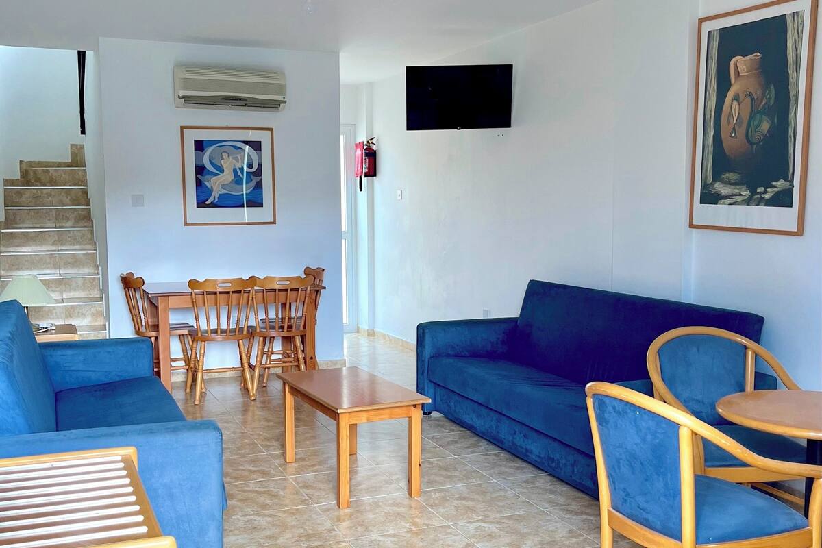 A living room with a blue sofa, wooden chairs, a small table, a TV mounted on the wall, and a staircase leading to another room.