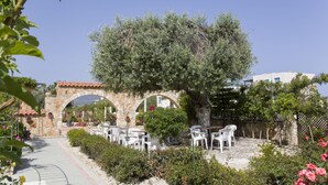 An outdoor seating area with white plastic chairs and tables, surrounded by greenery and a stone archway. - Eliofos Elegant Maisonettes (Polis)