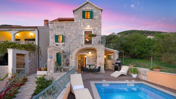 A stone building with a pool, outdoor seating, and a staircase leading to a balcony.