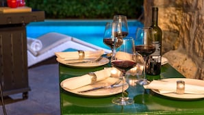 A dining table set with plates, cutlery, and wine glasses, with a bottle of wine and a pool in the background.