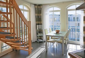 Une chambre d’hôtel moderne avec un escalier en bois, un coin repas comprenant une table et des chaises, une bibliothèque et de grandes fenêtres offrant une vue sur la ville.
