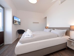 A hotel room with a large bed, a flat-screen TV mounted on the wall, a desk with a lamp, and a chair. - Ohlerich storage app. 27 (Wismar)