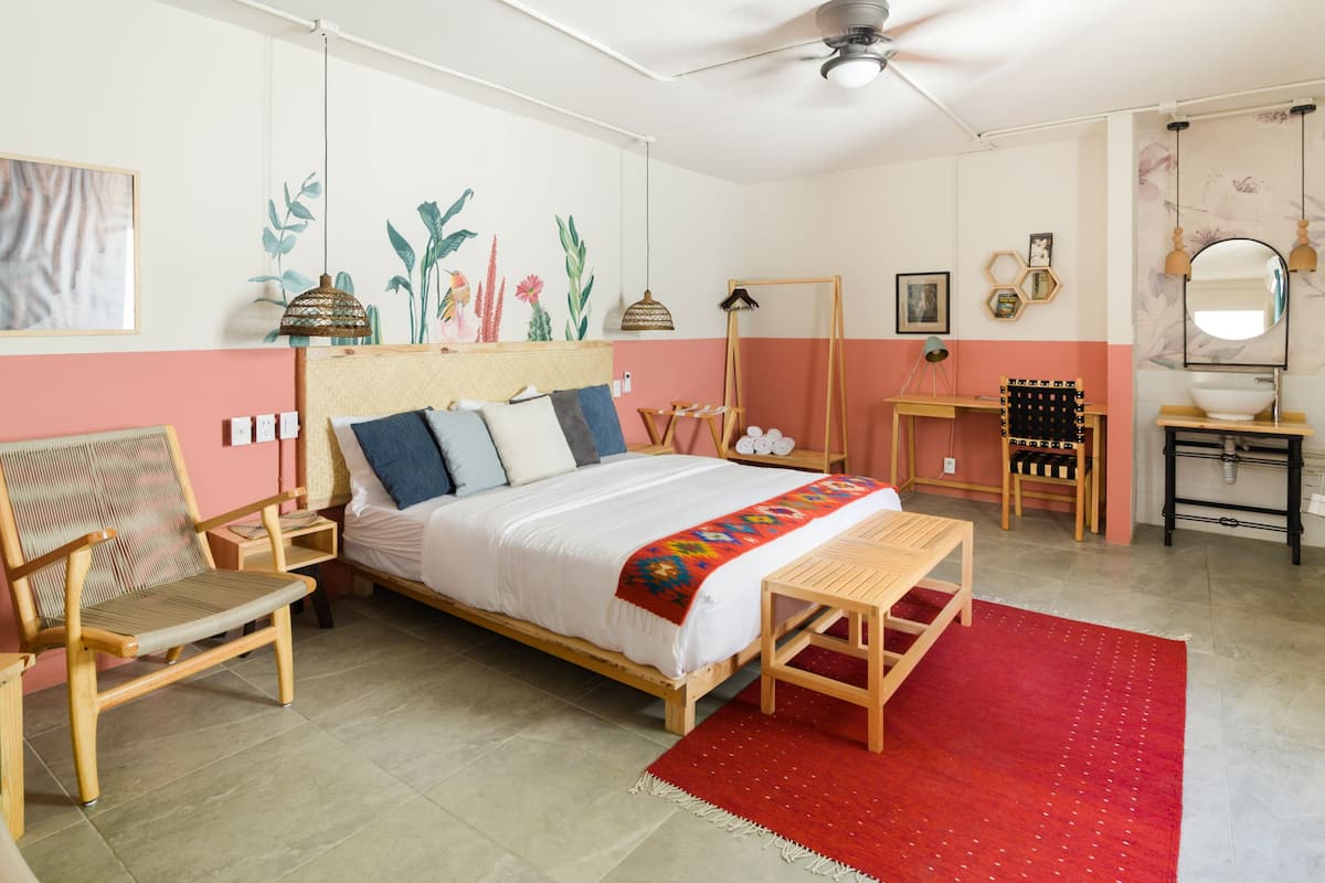 A bedroom with a bed, a wooden chair, a small table, a red rug, and a mirror.