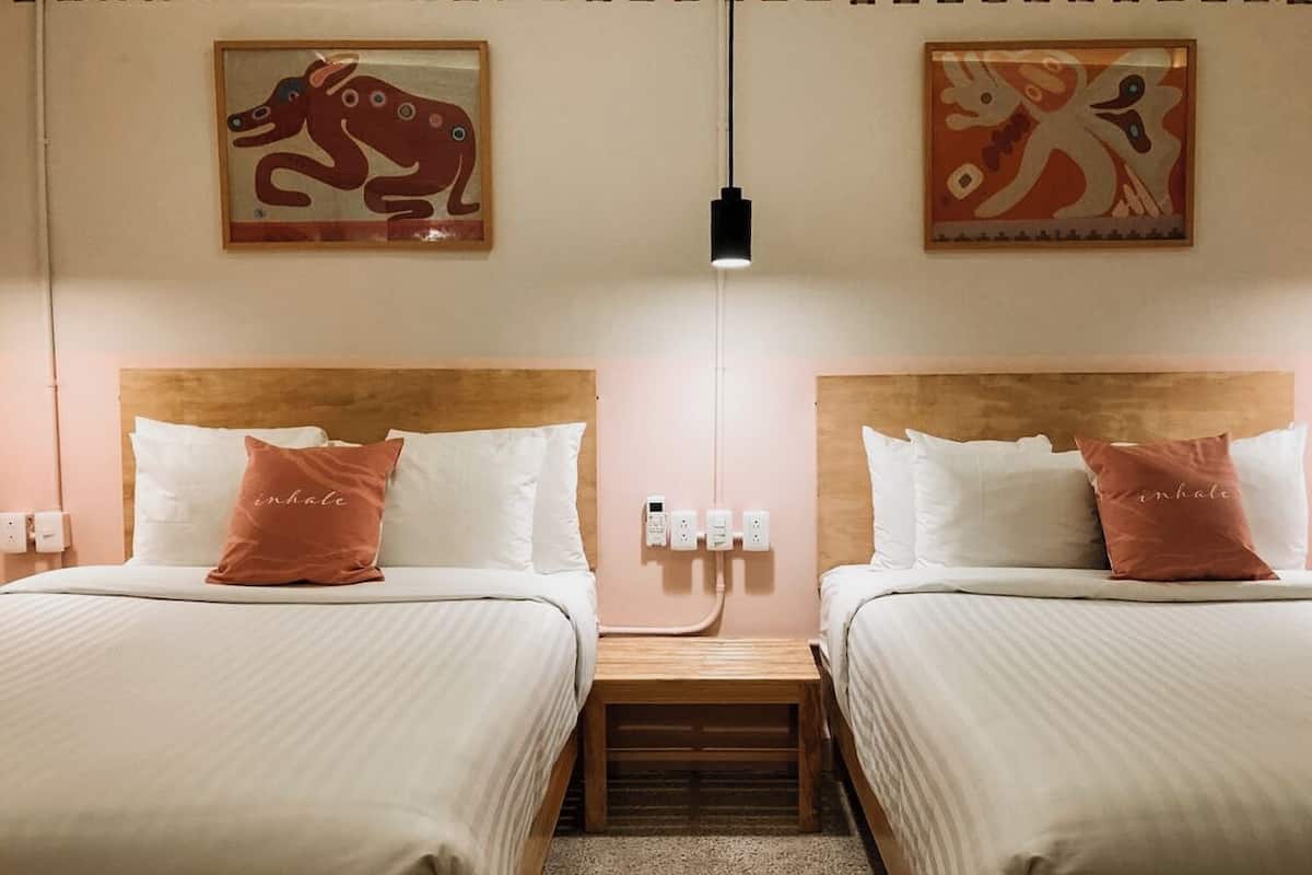 A hotel room with two beds, each with white bedding and a single rose gold pillow. There are wall-mounted artworks above the beds, a bedside table between the beds, and a single light fixture hanging from the ceiling.
