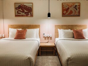 Habitación de hotel con dos camas, cada una con ropa de cama blanca y una única almohada color rosa dorado. Hay obras de arte colgadas en la pared sobre las camas, una mesita de noche entre ellas y una lámpara colgante del techo.