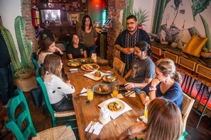 Un grupo de personas cenando en una mesa de madera en un restaurante con sillas verdes, decoración de cactus y una pared de ladrillo.