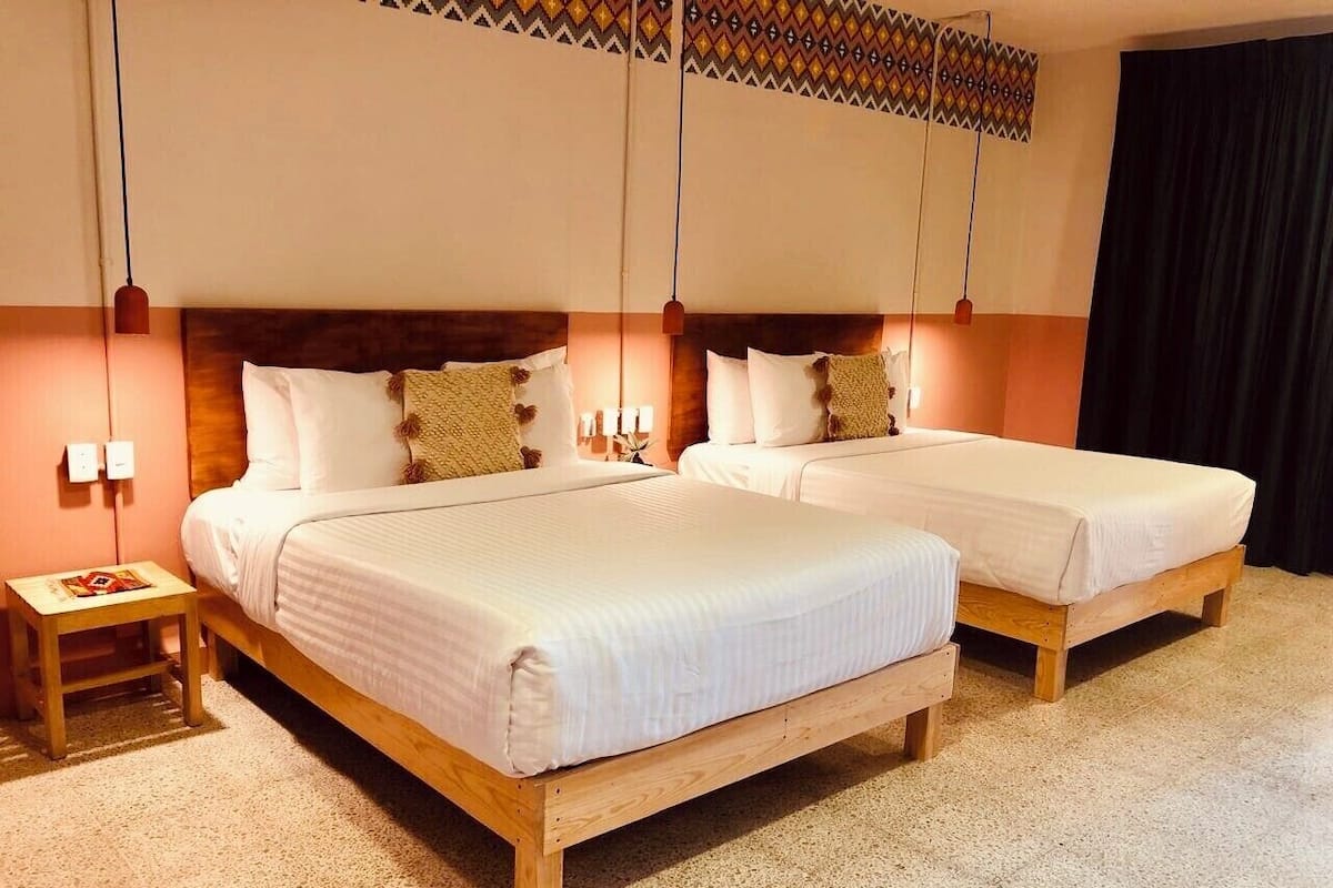 A hotel room with two wooden beds, each with white bedding and decorative pillows. There is a small bedside table with a lamp and a book. The room has a speckled floor and a patterned ceiling.
