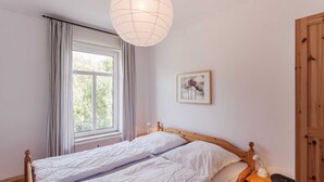 A bedroom with a wooden bed, a bedside table with a lamp, a window with curtains, and a wall-mounted picture. - Old village pitcher FW 3 - FW 3, north-east (Kronsgaard)