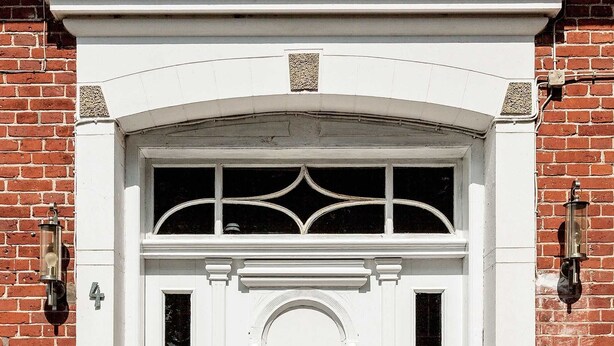 A white door with a decorative arch and two windows, flanked by brick walls and two wall-mounted lanterns. - Old village pitcher FW 3 - FW 3, north-east (Kronsgaard)