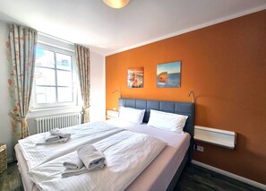 Una habitación de hotel con cama, ventana con cortinas, radiador y una obra de arte en la pared.