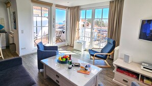 Un salon comprenant un canapé, un fauteuil, une table basse avec des fruits et des boissons, et un téléviseur posé sur un meuble.