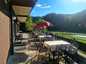 An outdoor patio with tables and chairs, a red umbrella, and a view of a river and forest.