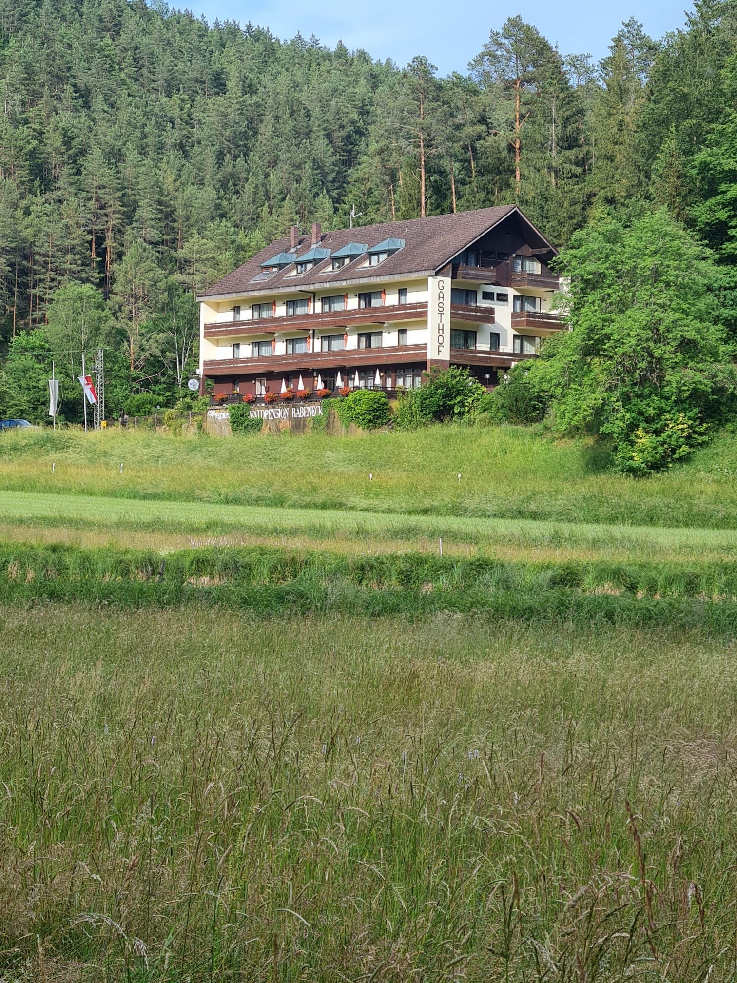 Ein mehrstöckiges Gebäude mit Balkonen, umgeben von einer grünen Wiese und bewaldeten Hügeln.