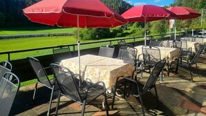Espace extérieur aménagé avec des parasols rouges, des tables recouvertes de nappes à motifs et une vue sur un champ verdoyant et une forêt.