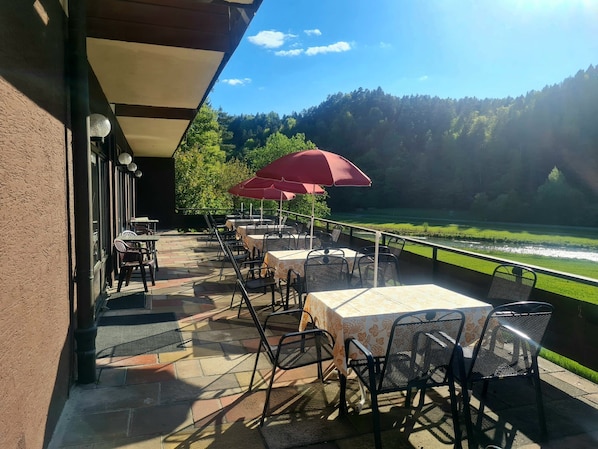 An outdoor patio with tables and chairs, a red umbrella, and a view of a river and forest.
