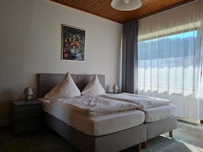 A bedroom with a bed, two bedside tables, a lamp, a painting, and a large window with curtains. - Double room no. 22 - Waldpension Rabeneck (Waischenfeld)