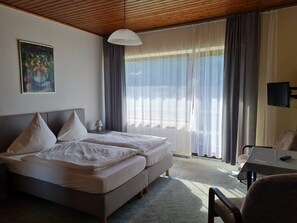 A bedroom with a bed, a desk, a chair, a TV, and a window with curtains.