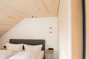 Une chambre avec un plafond en pente, un lit avec une literie blanche, un petit tabouret en bois et un interrupteur fixé au mur.