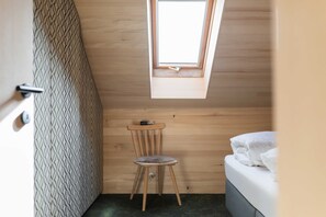 Ein kleiner Raum mit schrägem Dach, einem Oberlicht, einem Holzstuhl, einem Bett und einer Tür.