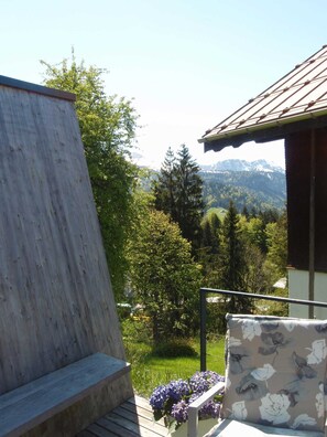 Une terrasse en bois avec un banc, un coussin à motifs floraux et une vue sur un paysage montagneux.