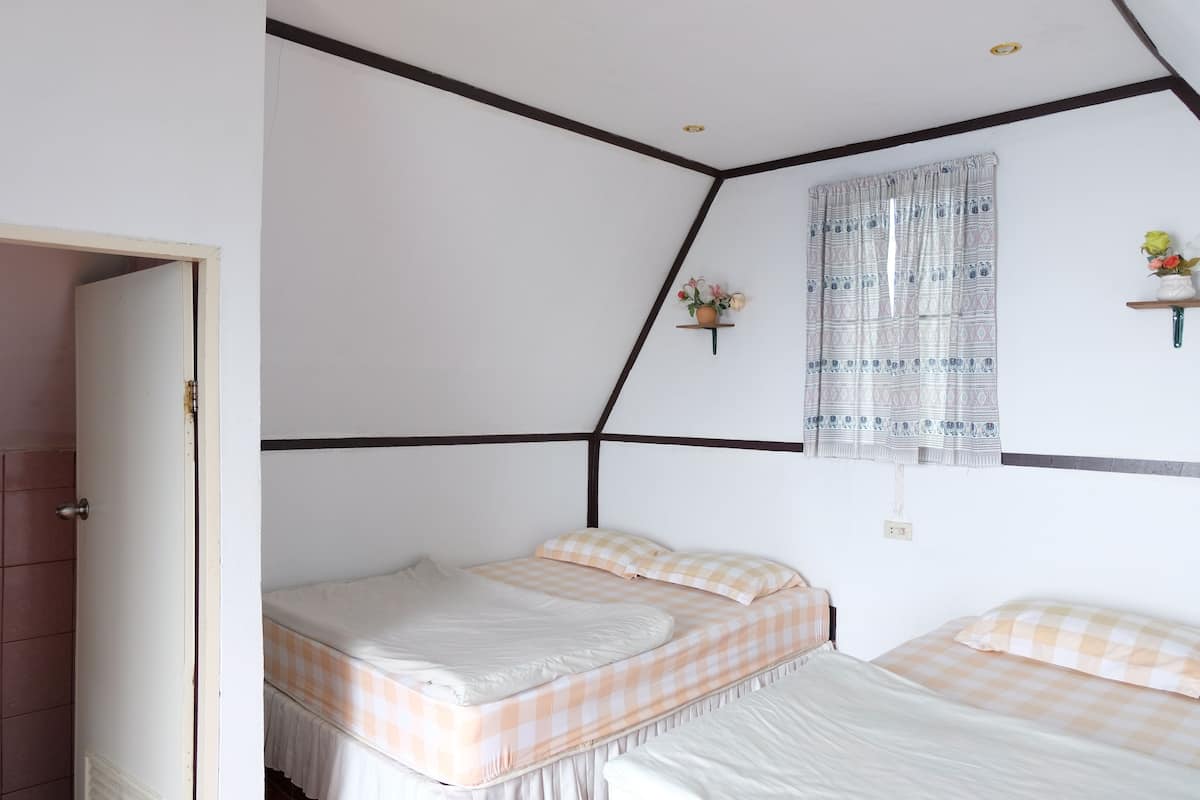 A bedroom with two beds, a window with patterned curtains, and a small shelf with flowers.