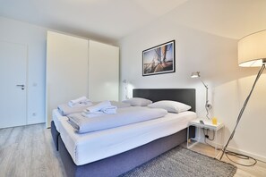 Cama bem arrumada com lençóis brancos, cabeceira cinza e uma pequena mesa de cabeceira com abajur e controle remoto.