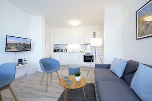 Uma sala de estar moderna com um sofá cinza, uma poltrona azul e uma pequena mesa de centro de madeira. Uma cozinha visível ao fundo apresenta armários brancos e um forno embutido.