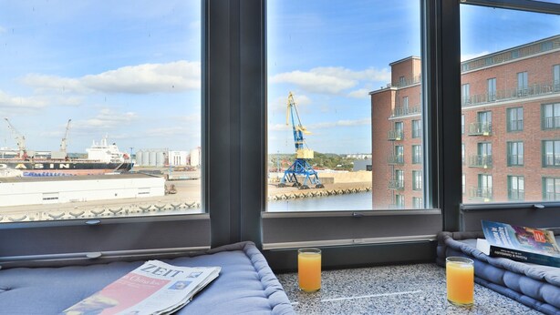Une chambre d’hôtel avec une grande fenêtre donnant sur un chantier de construction, un journal et deux verres de jus d’orange posés sur le rebord de la fenêtre.