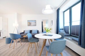 Uma sala de estar moderna com uma mesa redonda, cadeiras azuis, um sofá cinza e uma janela grande com persianas.
