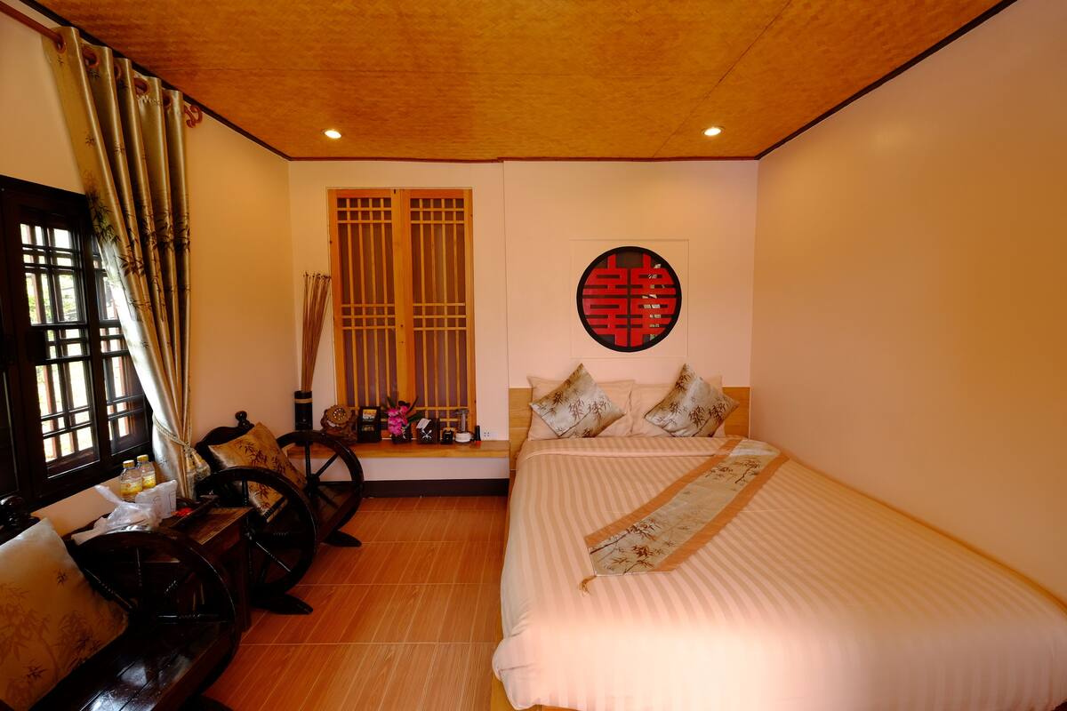 Una habitación con una cama, un banco de madera, un ventanal con cortinas de bambú y una pared con un diseño circular rojo.