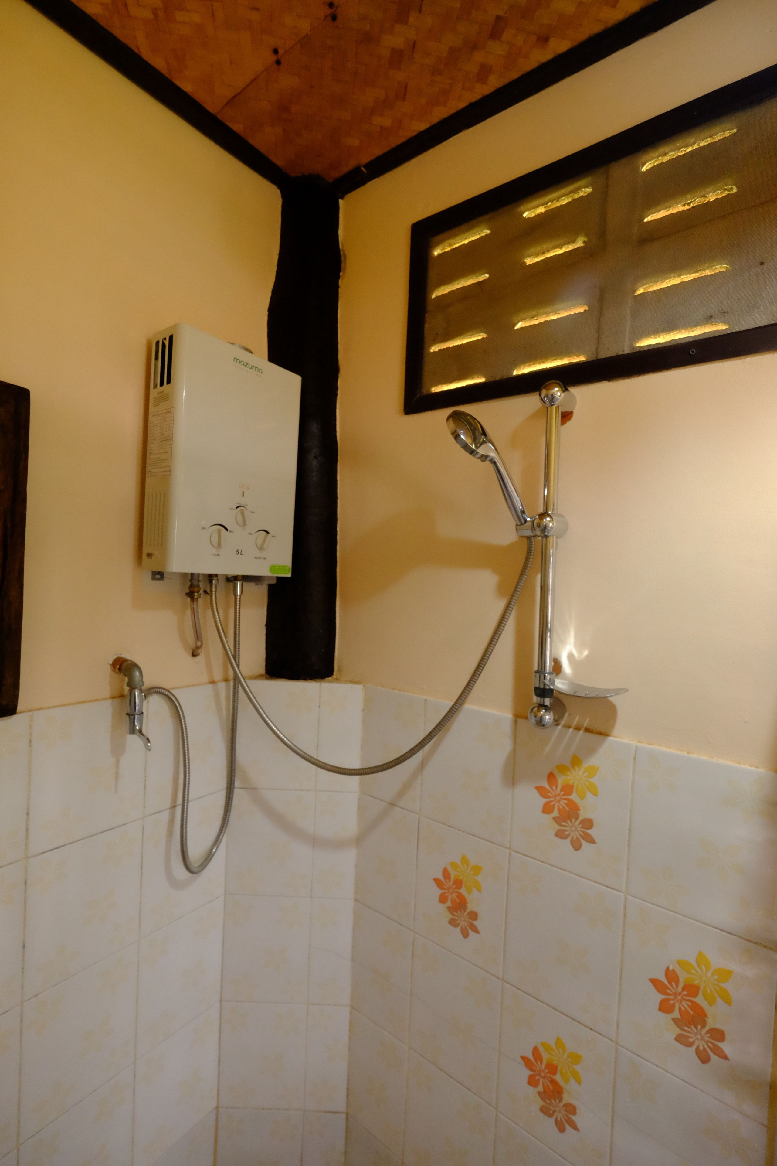 A bathroom with a shower, a water heater, and a tiled wall with floral patterns.