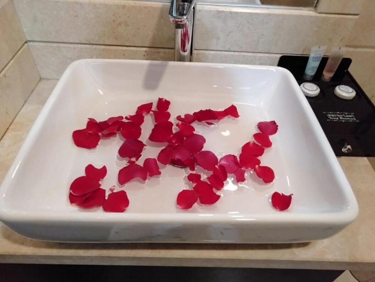 A white rectangular sink with red rose petals floating in the water, a faucet, and toiletries on a black tray.