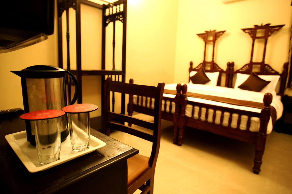 A room with a bed, a wooden chair, a table with two glasses and a kettle, and a wall-mounted TV.