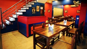 A restaurant with a red and blue color scheme, wooden tables and chairs, a staircase, and a large screen on the wall. - Hotel Ajit Mansion (Jodhpur)