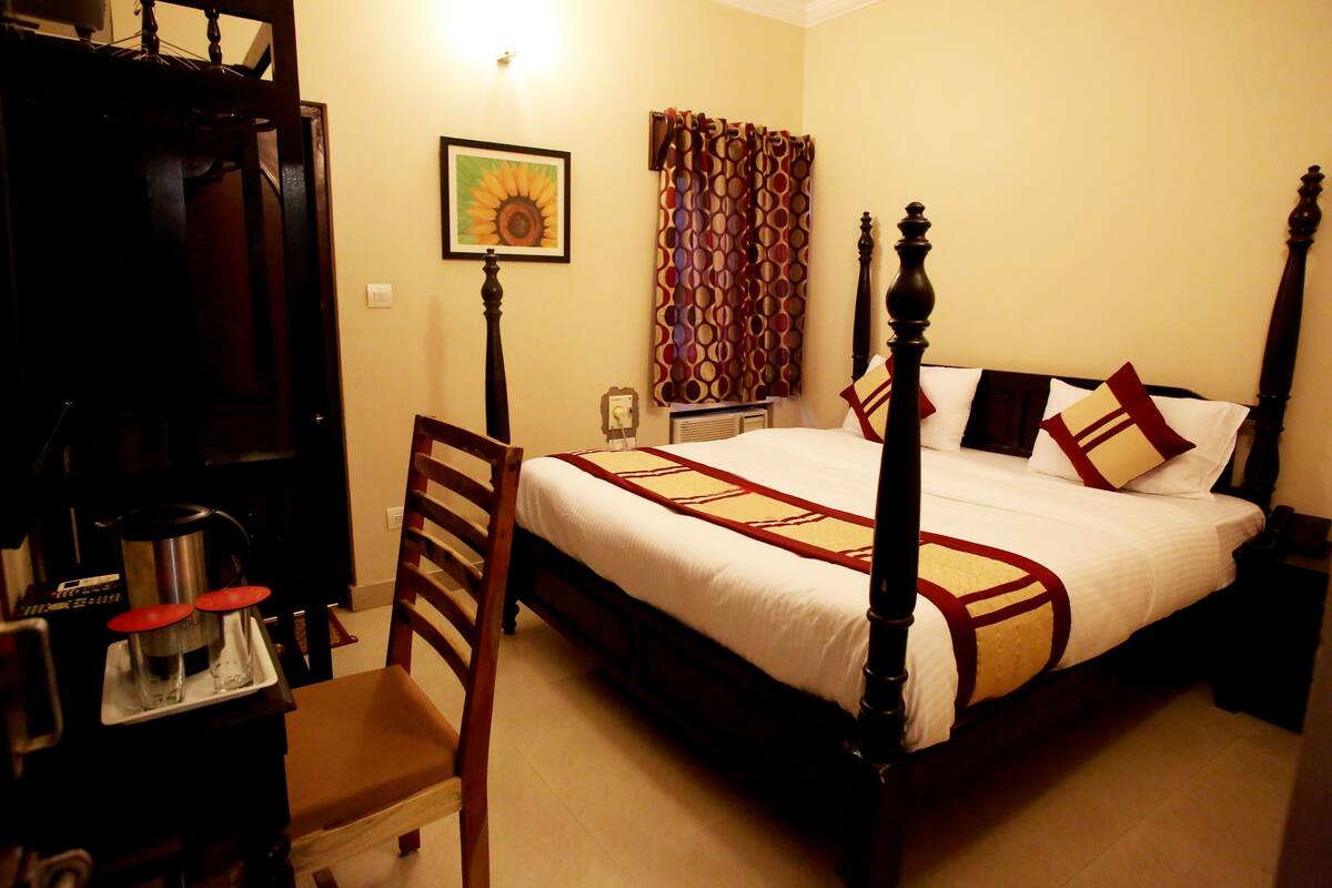 A hotel room with a four-poster bed, a wooden chair, a desk with a kettle and glasses, and a painting on the wall.