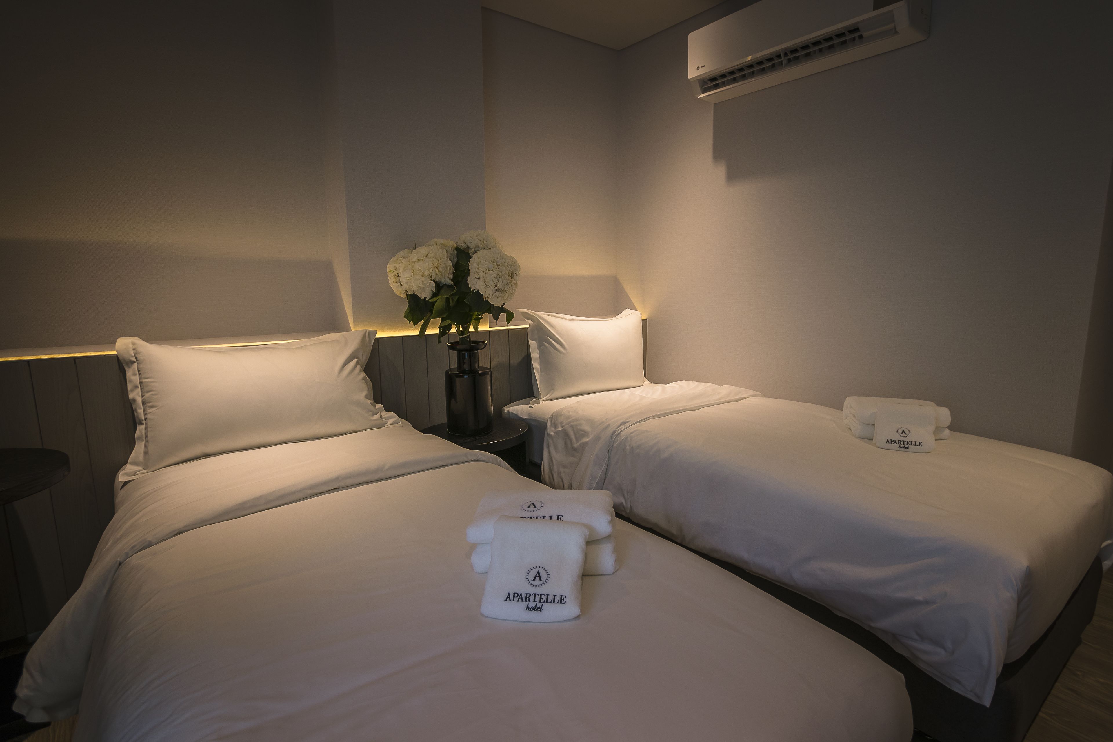 A hotel room with two beds, each with a white pillow and a towel with the hotel name. There is a vase with white flowers on the nightstand between the beds.