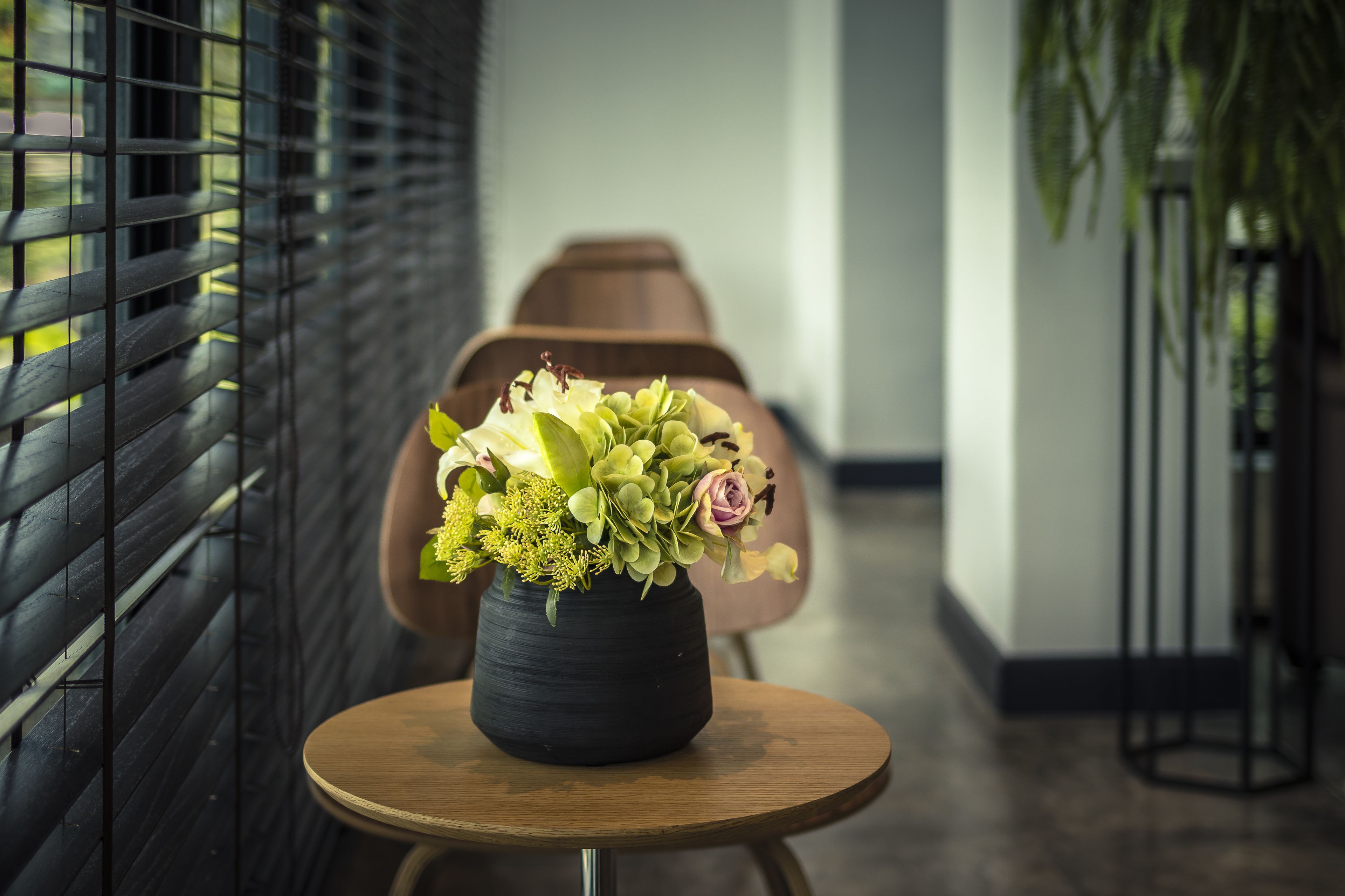 Et moderne interiør med et rundt træbord, der bærer en sort vase med blomster, en sort metalstol og en plante i baggrunden.