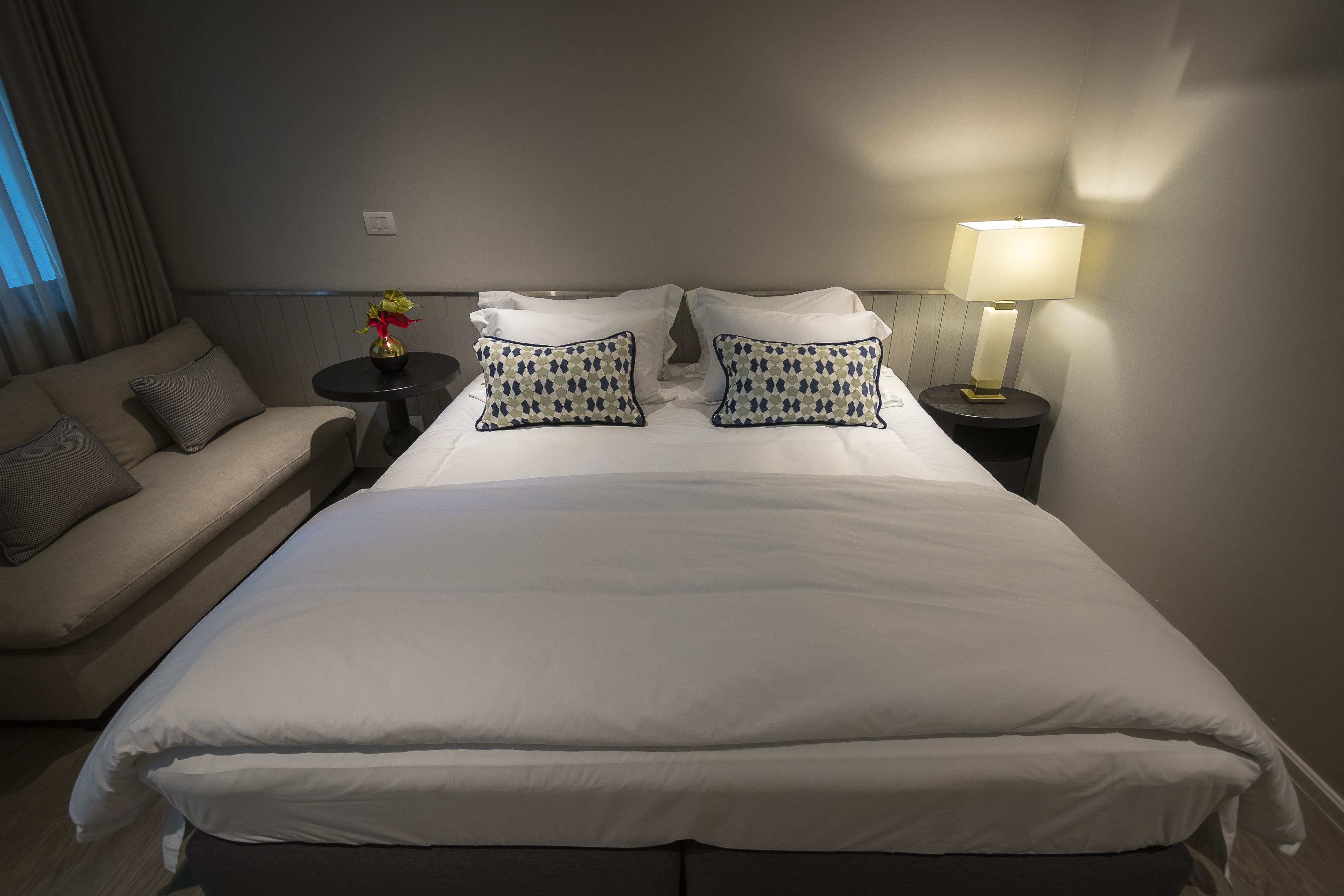 A neatly made bed with white linens, two decorative pillows, a bedside table with a lamp, and a sofa with cushions.