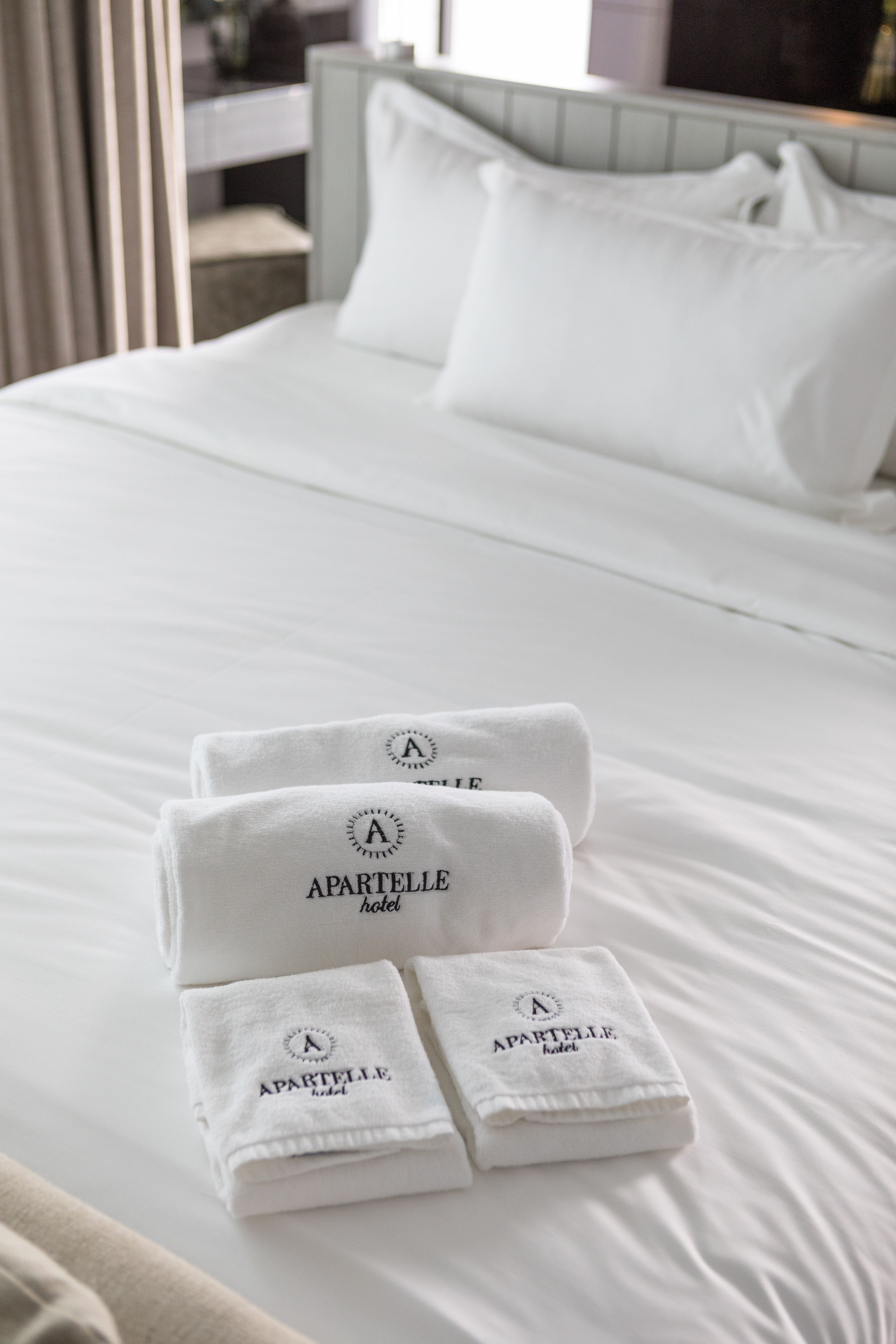 Un lit bien fait, avec du linge de lit blanc et trois serviettes pliées sur lesquelles on peut lire le texte « APARTELLE hotel ».