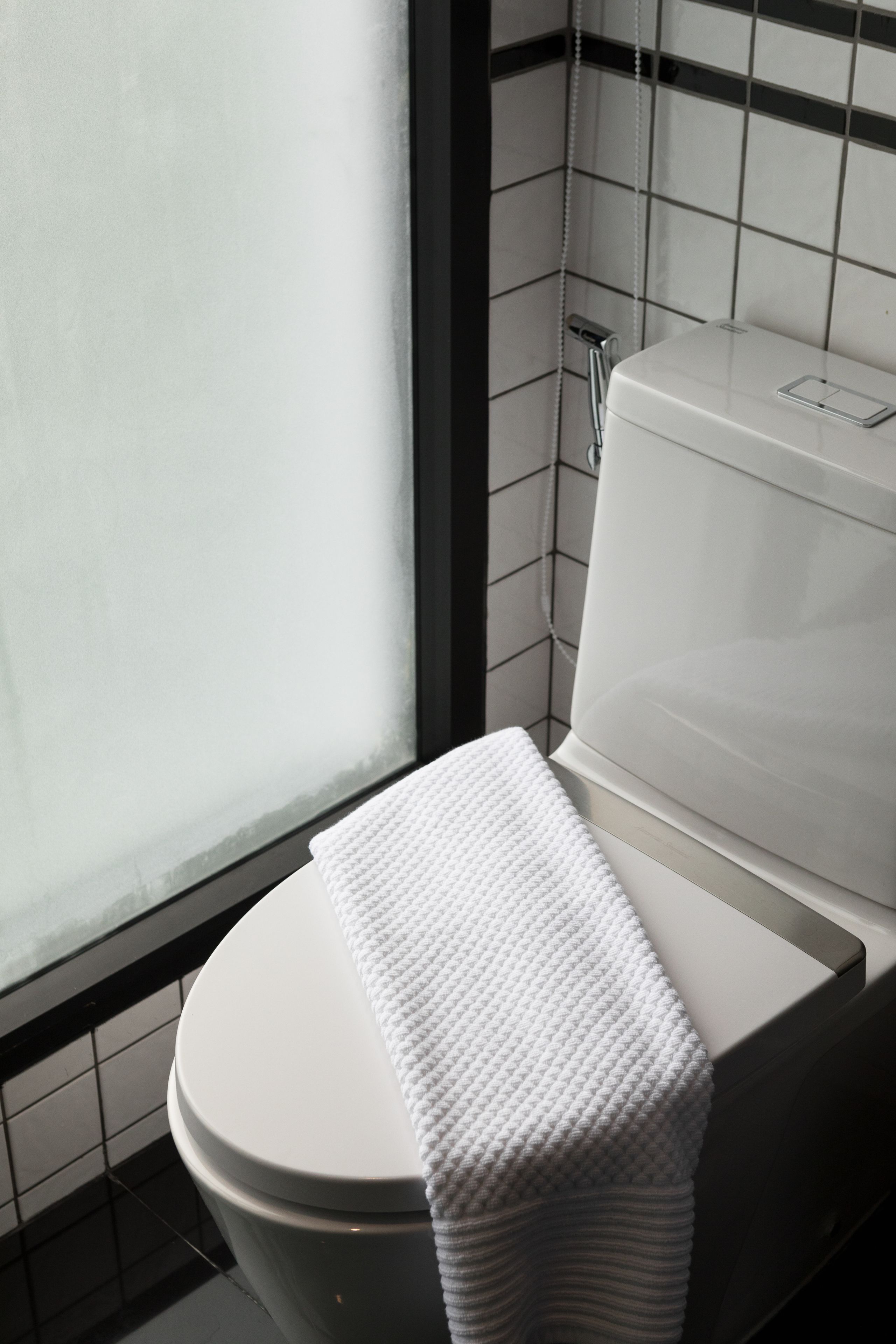 A white toilet with a white towel on the seat, a visible flush handle, and a tiled wall with a black border.