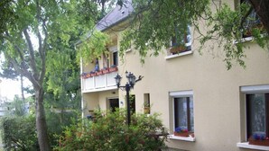 Un bâtiment à deux étages doté d’un balcon, de fenêtres ornées de jardinières et d’un espace jardin avec un arbre et des arbustes.