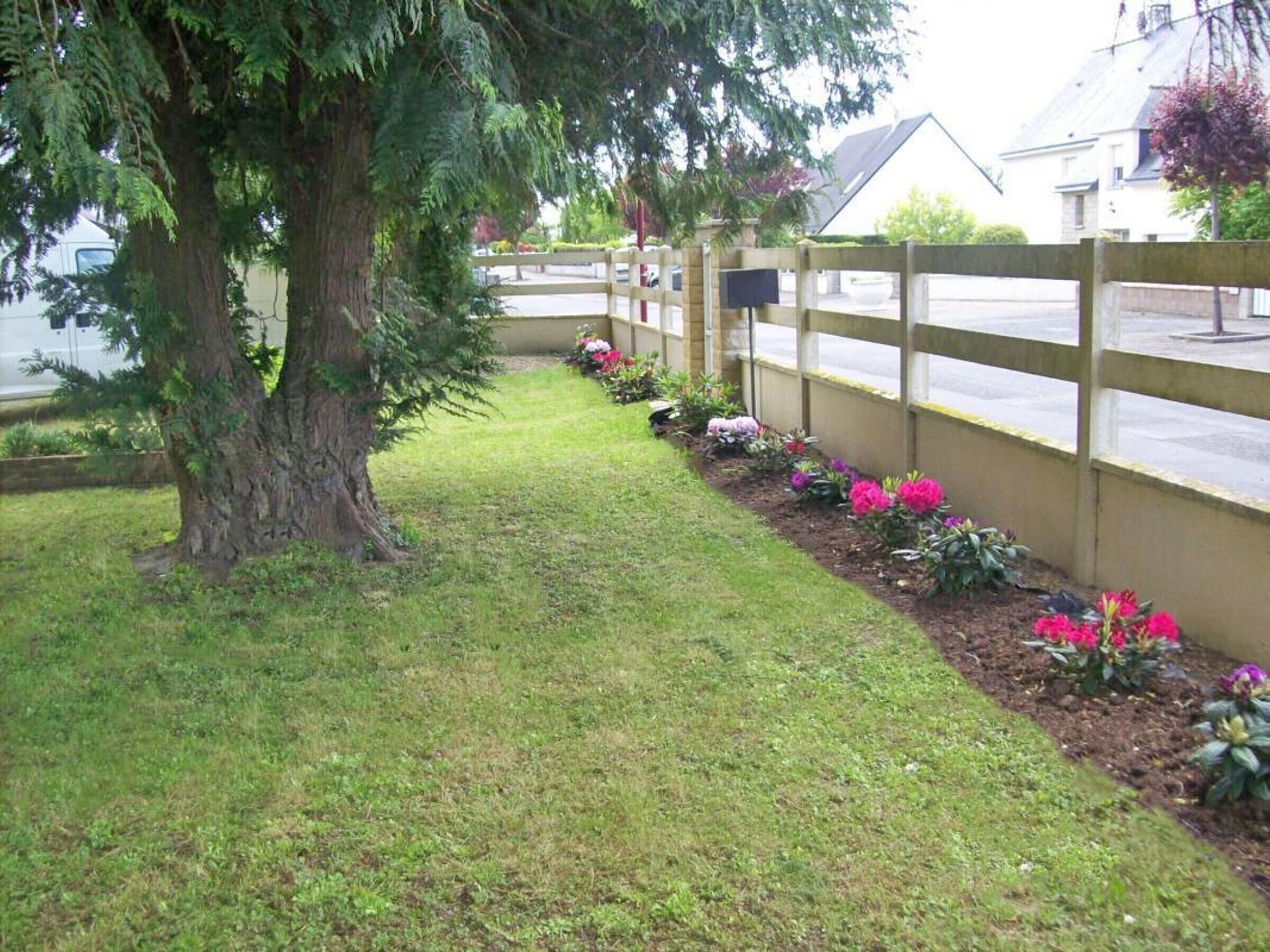 A pathway lined with a wooden fence and flowering plants, leading to a residential area with houses and a parked vehicle.