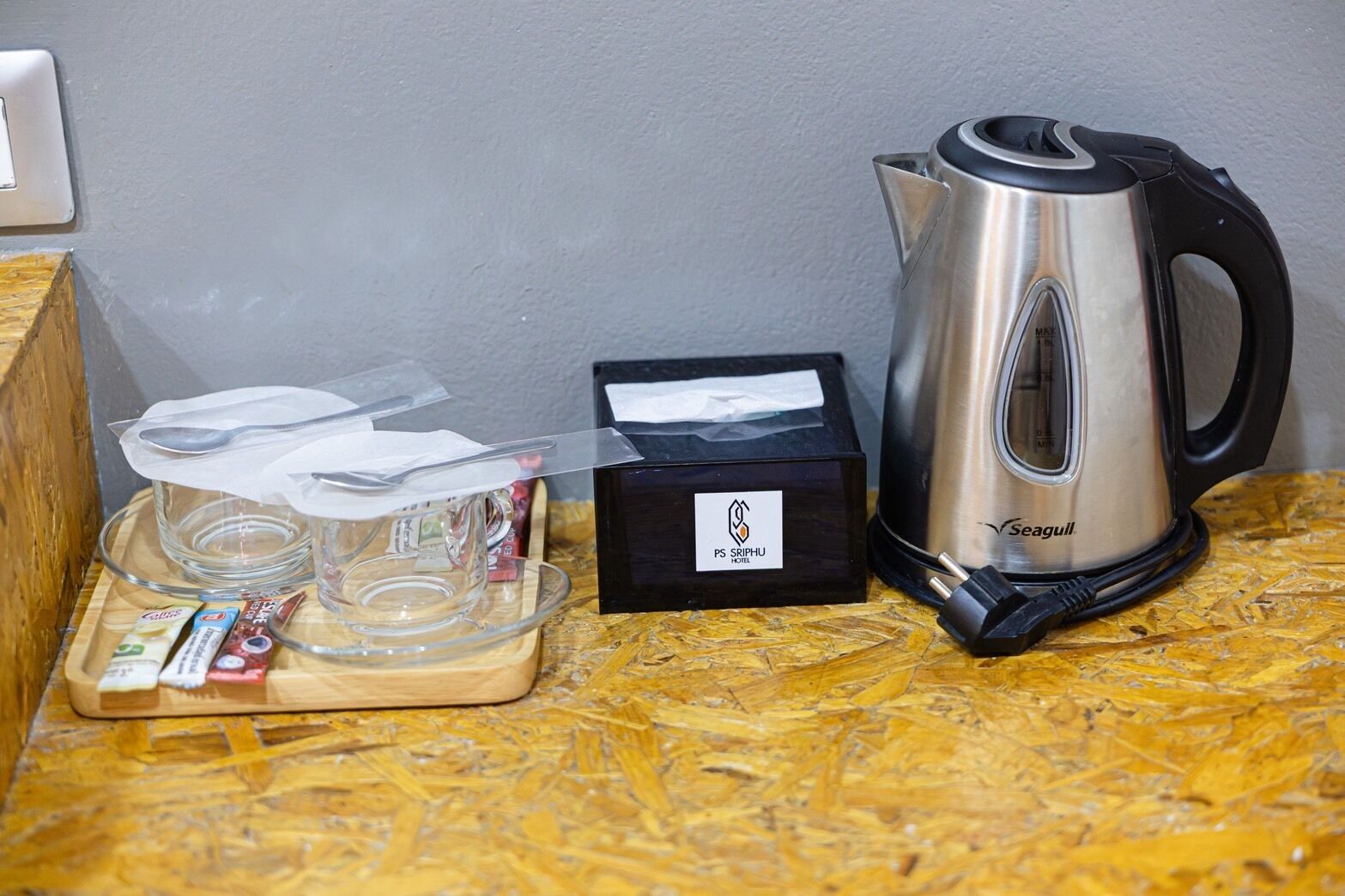 Eine Seagull-Kaffeemaschine, eine Taschentuchbox und ein Tablett mit Gläsern und einem Löffel auf einer Holztischplatte.