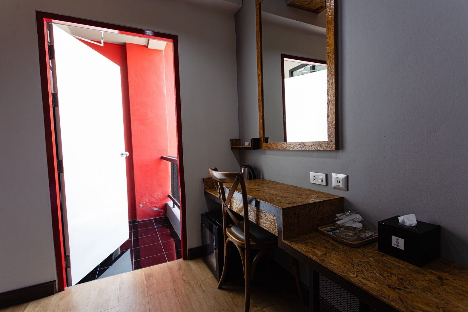 A hotel room with a wooden desk, a chair, a mirror, and a red door.