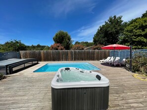 Un patio trasero con piscina, jacuzzi y sillones de jardín bajo un paraguas rojo.