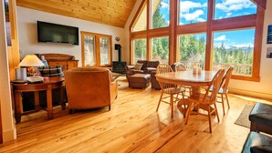 Amplia sala de estar con techo de madera, ventanas grandes que ofrecen vista a la montaña, chimenea y televisor montado en la pared.