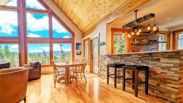 Una cocina amplia con salpicadero de piedra, mesa de comedor de madera y taburetes de bar. Grandes ventanales ofrecen vistas a las montañas.
