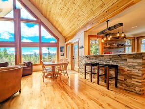A spacious kitchen with a stone backsplash, wooden dining table, and bar stools. Large windows offer a view of the mountains. - Leavenworth Private 5 acre Mountain Vista Retreat (Leavenworth)